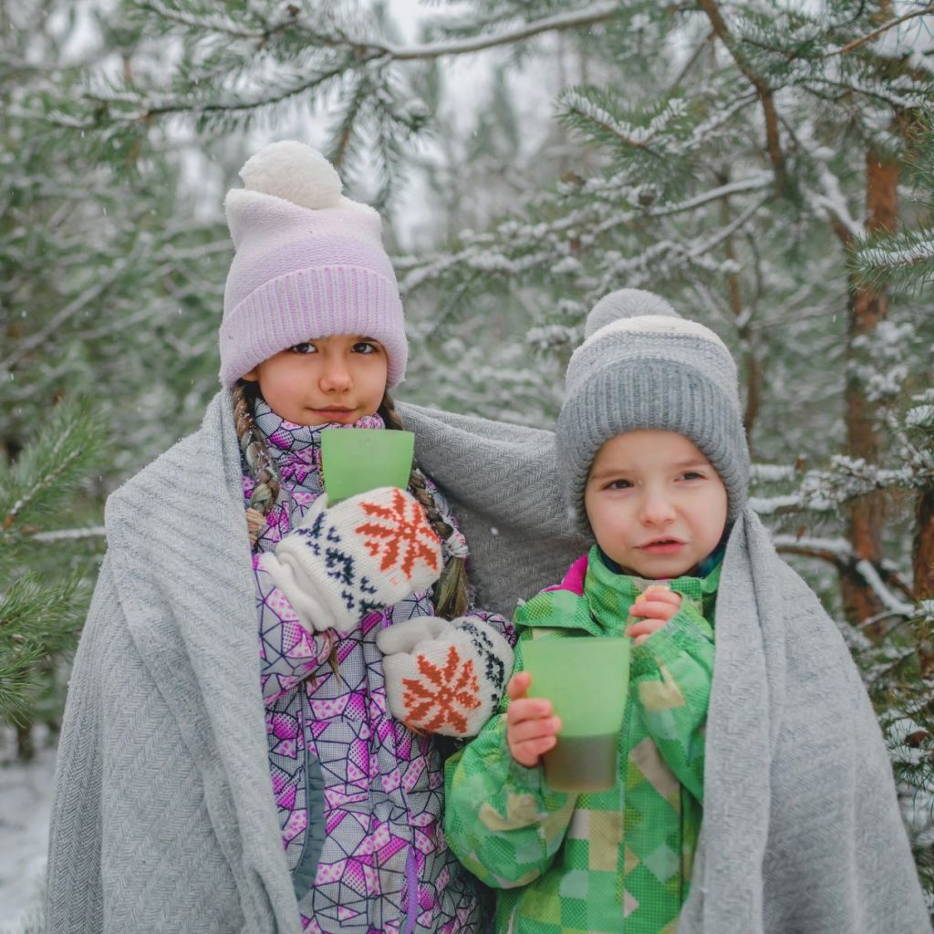 two kids holding drinks in a snowy woods wrapped in blankets wearing hats and coats