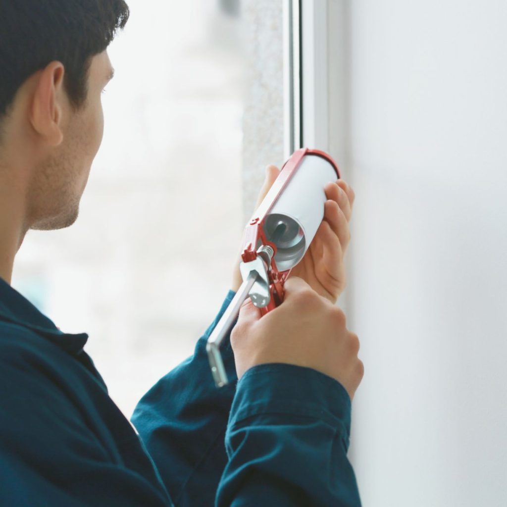 man in blue shirt using a caulk gun to seal windows